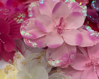 May include: A collection of artificial fabric flowers in shades of pink, white, and yellow. Each petal is edged with iridescent, shimmering sequins that catch the light. The centers of the flowers feature small, delicate beads in matching tones.