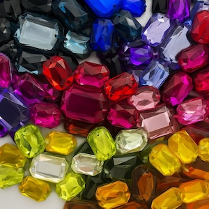 May include: A collection of faceted, rectangular gemstones in a variety of colors. The gemstones range from black, blue, and purple to red, pink, yellow, and orange. The gems are arranged in a gradient pattern.
