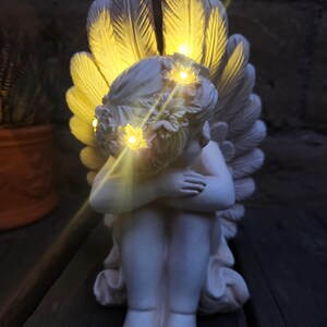 May include: A white cherub statue with illuminated wings and a floral crown. The angel is seated with its arms wrapped around its knees. The wings and crown have small, warm-toned lights. The statue is a decorative item.