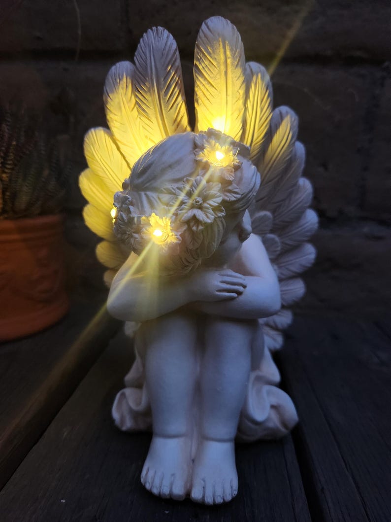 May include: A white cherub statue with illuminated wings and a floral crown. The angel is seated with its arms wrapped around its knees. The wings and crown have small, warm-toned lights. The statue is a decorative item.