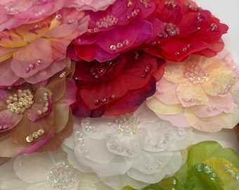 May include: A collection of layered fabric flowers in various colors including pink, red, white, yellow, brown, and green. Each flower features delicate, translucent petals adorned with small, iridescent beads scattered across the surface.