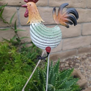 May include: A metal rooster garden stake with a red comb, green and white body, and black tail feathers. The rooster is standing on a thin metal stake.