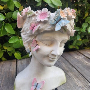 May include: A white, sculpted head planter adorned with pink and blue butterflies and flowers. The planter has a serene expression and is set against a backdrop of green foliage and wooden planks.