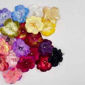 May include: A collection of layered fabric flowers in various colors including blue, white, yellow, green, purple, pink, red, and black. Each flower features delicate petals and a small, sparkling rhinestone or beaded center detail.