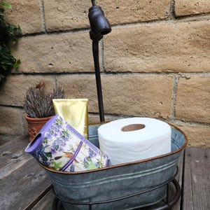 May include: A galvanized metal tub-shaped bathroom organizer with a faucet-shaped handle. The tub holds a roll of toilet paper, a decorative container, and a gold tube. The organizer sits on a dark metal stand.