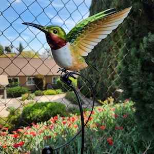 May include: A vibrant metal hummingbird sculpture with open wings, mounted on a black, curved stand. The bird displays green, red, white, and gold colours. The background includes a chain-link fence, greenery, and a blue sky.