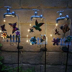 May include: Three solar-powered garden stakes with metal water faucets, each with a different metal animal and flowers. The stakes have a white picket fence design and are illuminated with warm white fairy lights.