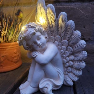 May include: A white cherub statue with feathered wings, illuminated by warm yellow lights. The cherub is seated, with its head resting on its knees. The statue is made of a smooth material and is placed on a wooden surface.