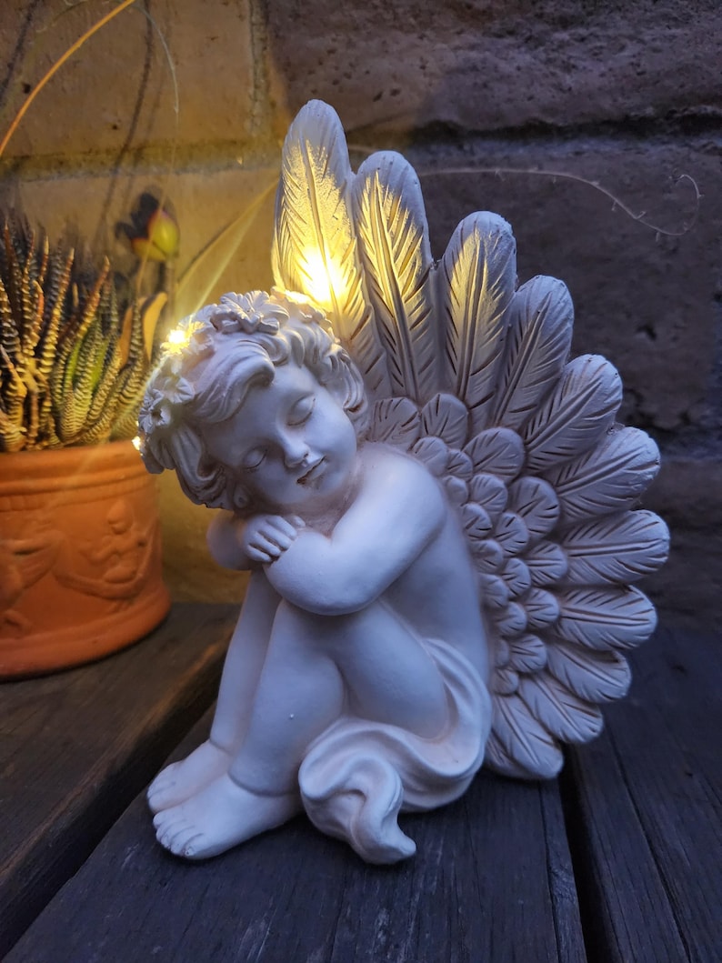 May include: A white cherub statue with feathered wings, illuminated by warm yellow lights. The cherub is seated, with its head resting on its knees. The statue is made of a smooth material and is placed on a wooden surface.
