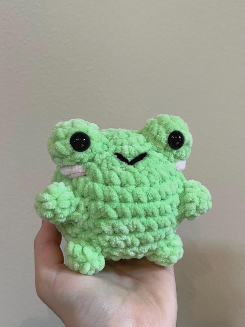 May include: A small green crocheted frog plush toy with black eyes and a pink mouth. The frog is sitting in a person's hand.