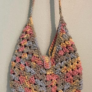 Puede incluir: Bolso de hombro de ganchillo hecho a mano con un patr&oacute;n de cuadrados de la abuela. El bolso presenta una mezcla de colores pastel, incluyendo rosa, amarillo, gris y beige. El bolso tiene un cierre de solapa y dos correas para el hombro.
