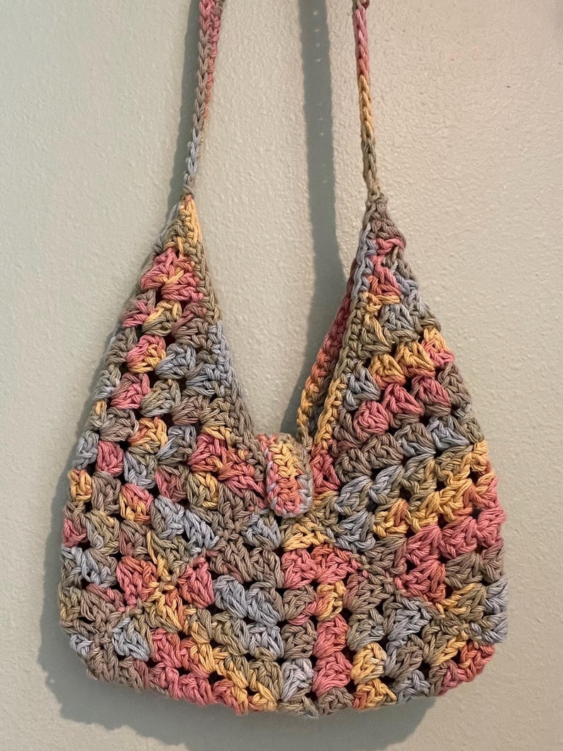 Puede incluir: Bolso de hombro de ganchillo hecho a mano con un patr&oacute;n de cuadrados de la abuela. El bolso presenta una mezcla de colores pastel, incluyendo rosa, amarillo, gris y beige. El bolso tiene un cierre de solapa y dos correas para el hombro.
