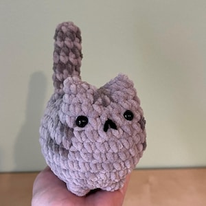 May include: A handmade, plush cat toy in shades of gray. The cat has a round body, small ears, and a tail. Black safety eyes and a small nose complete the face. The toy is made of a soft, textured yarn.