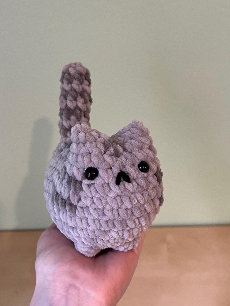 May include: A handmade, plush cat toy in shades of gray. The cat has a round body, small ears, and a tail. Black safety eyes and a small nose complete the face. The toy is made of a soft, textured yarn.