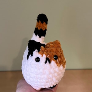 May include: A handmade, plush cat toy in white, orange, black, and brown. The cat has a round body, black button eyes, and a small, upturned tail. The cat's face is a mix of orange and white, with black accents.