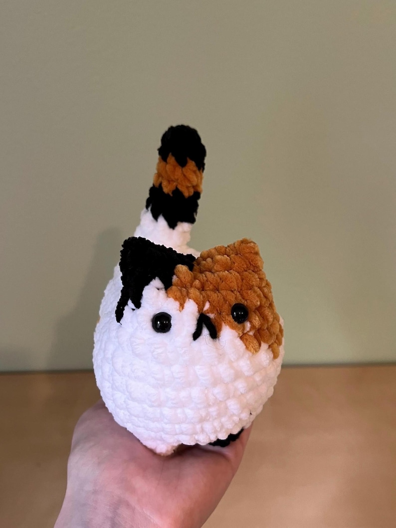 May include: A handmade, plush cat toy in white, orange, black, and brown. The cat has a round body, black button eyes, and a small, upturned tail. The cat's face is a mix of orange and white, with black accents.
