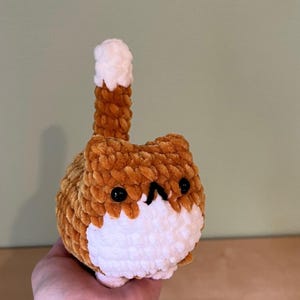 May include: A handmade, plush cat toy in shades of brown and white. The cat has a round body, a white face, and black eyes. Its tail is upright, with a brown base and a white tip. The toy is made of soft, textured yarn.