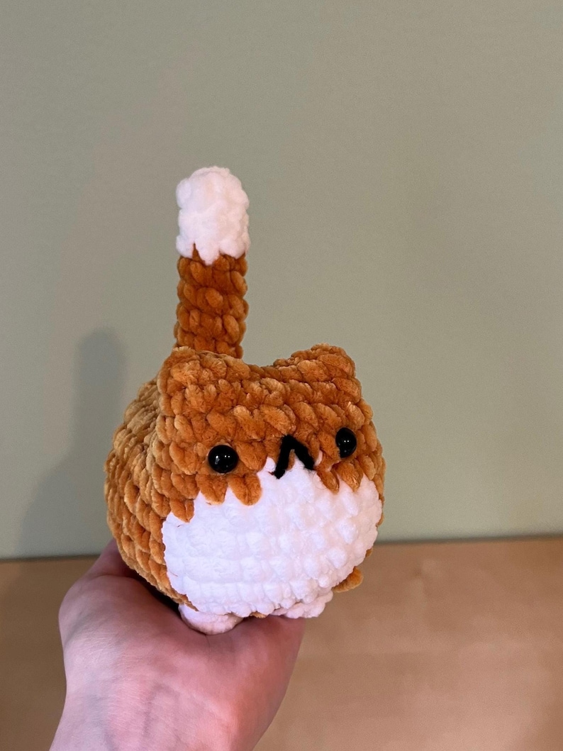 May include: A handmade, plush cat toy in shades of brown and white. The cat has a round body, a white face, and black eyes. Its tail is upright, with a brown base and a white tip. The toy is made of soft, textured yarn.
