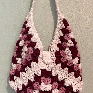 Puede incluir: Un bolso de crochet hecho a mano con un dise&ntilde;o de cuadrados de la abuela. El bolso presenta una combinaci&oacute;n de cuadrados blancos, burdeos y rosa claro. Tiene una larga correa blanca y un cierre de bot&oacute;n. El bolso est&aacute; dise&ntilde;ado para llevarse al hombro.