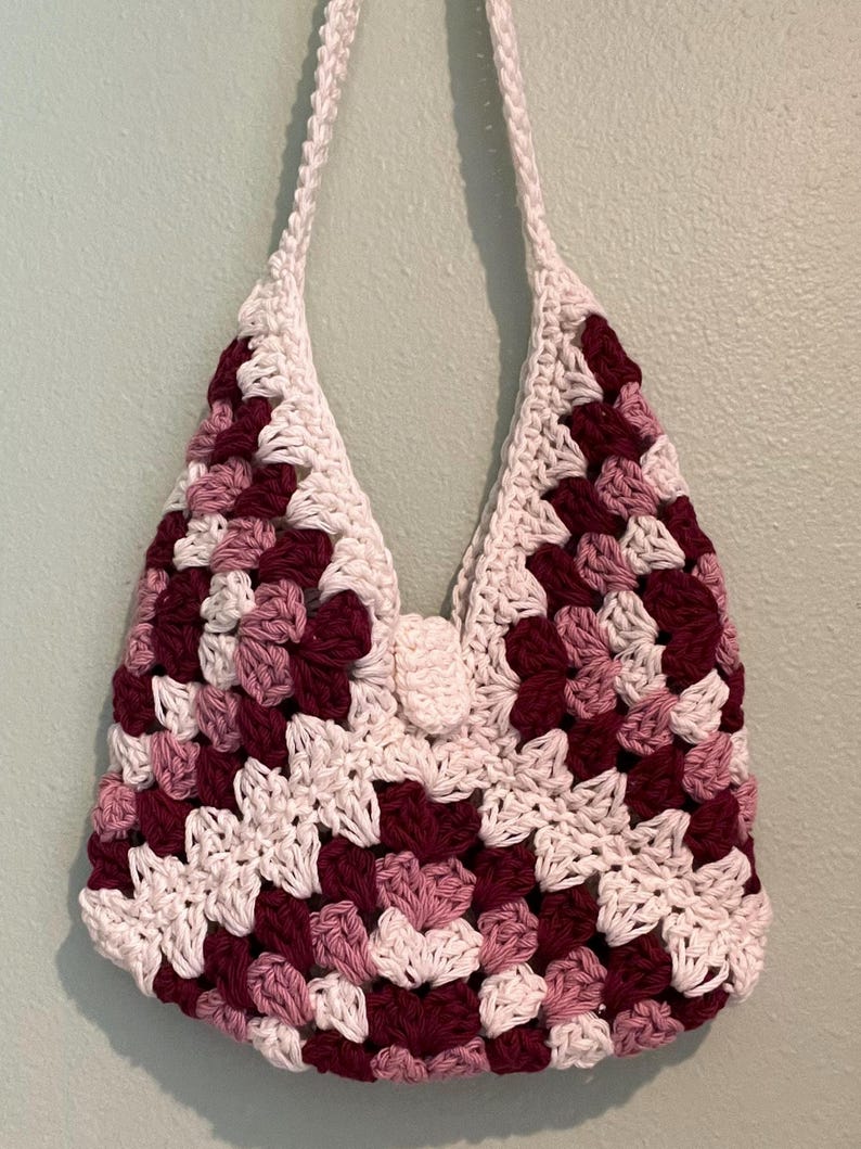Puede incluir: Un bolso de crochet hecho a mano con un dise&ntilde;o de cuadrados de la abuela. El bolso presenta una combinaci&oacute;n de cuadrados blancos, burdeos y rosa claro. Tiene una larga correa blanca y un cierre de bot&oacute;n. El bolso est&aacute; dise&ntilde;ado para llevarse al hombro.