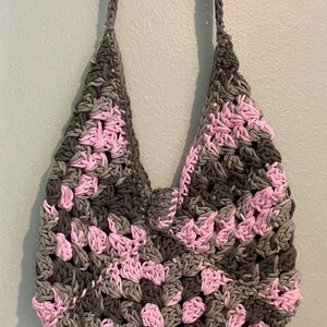 Puede incluir: Un bolso tote de crochet hecho a mano con un patr&oacute;n de granny square. El bolso presenta una combinaci&oacute;n de colores gris, rosa y gris oscuro. El bolso tiene dos correas y est&aacute; colgado contra una pared azul claro.
