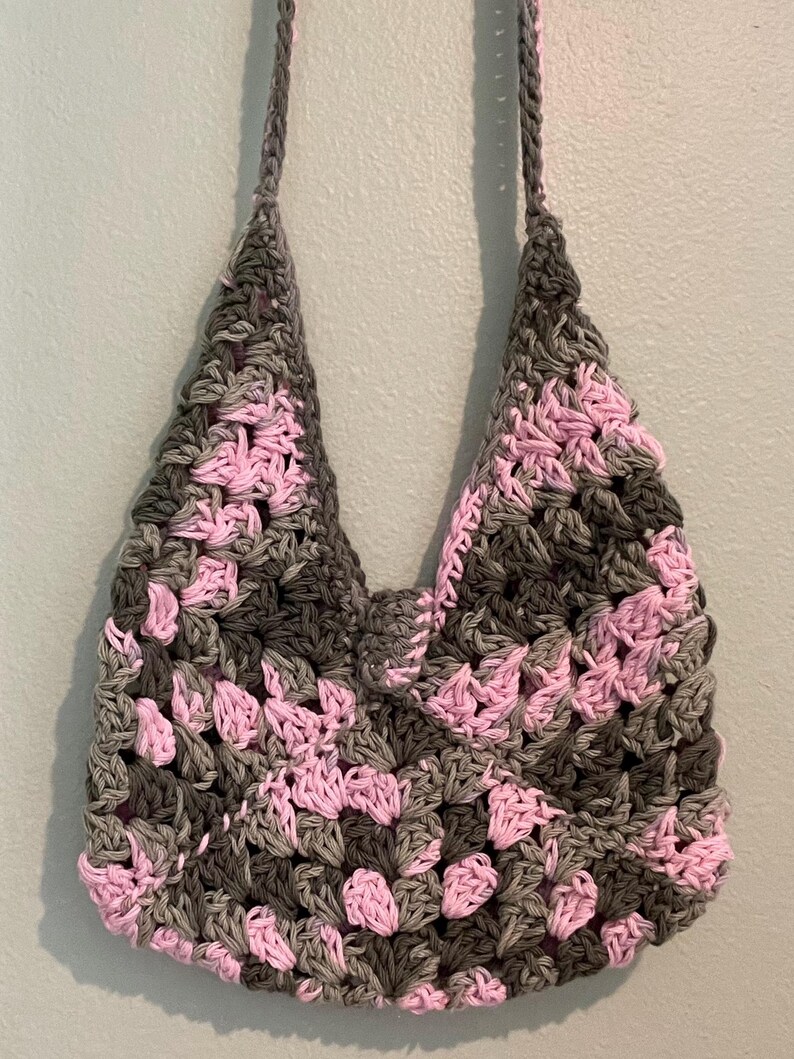 Puede incluir: Un bolso tote de crochet hecho a mano con un patr&oacute;n de granny square. El bolso presenta una combinaci&oacute;n de colores gris, rosa y gris oscuro. El bolso tiene dos correas y est&aacute; colgado contra una pared azul claro.