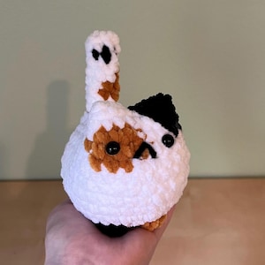 May include: A handmade, crocheted cat toy in white, brown, and black. The cat has a round body, a small tail, and black button eyes. The cat's head has a black patch and brown spots. The toy is held in a hand.