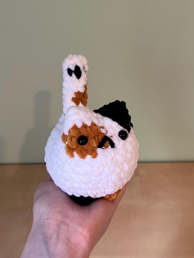 May include: A handmade, crocheted cat toy in white, brown, and black. The cat has a round body, a small tail, and black button eyes. The cat's head has a black patch and brown spots. The toy is held in a hand.