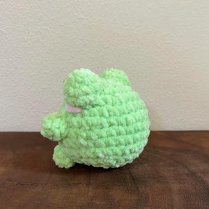 May include: A green crocheted frog plush toy with a pink mouth. The frog is sitting on a wooden surface.