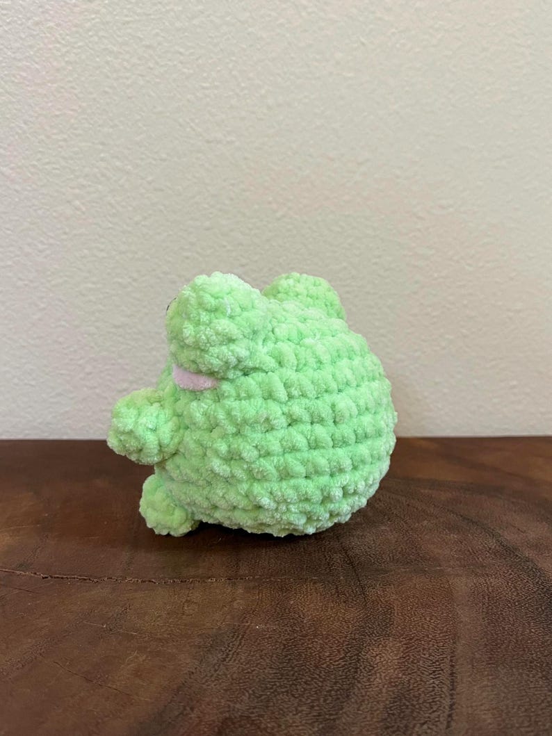 May include: A green crocheted frog plush toy with a pink mouth. The frog is sitting on a wooden surface.