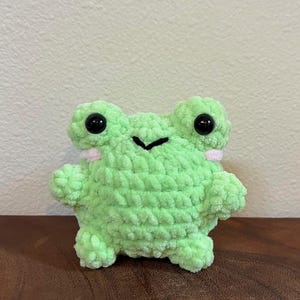 May include: A small green crocheted frog plush toy with black eyes and pink accents. The frog is sitting on a wooden surface.