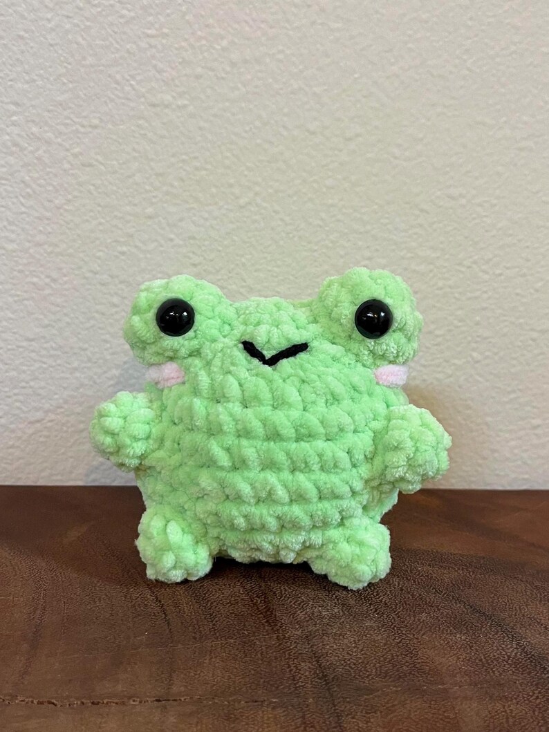 May include: A small green crocheted frog plush toy with black eyes and pink accents. The frog is sitting on a wooden surface.