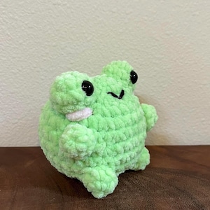 May include: A green crocheted frog plush toy with black eyes. The frog is sitting on a wooden surface.