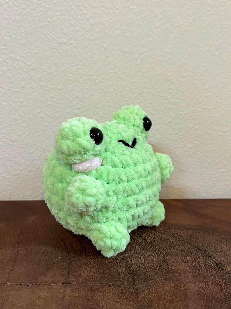 May include: A green crocheted frog plush toy with black eyes. The frog is sitting on a wooden surface.