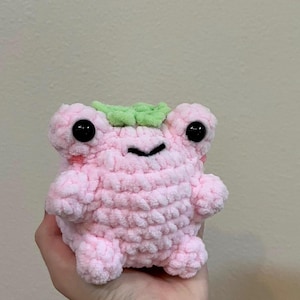 Puede incluir: Un juguete de peluche de rana de crochet rosa con ojos negros y una hoja de crochet verde en la cabeza.