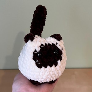 May include: A handmade crochet cat toy in white and brown. The cat has dark brown markings around the eyes, ears, and paws, with a matching tail. The toy is round and fluffy, with black safety eyes.