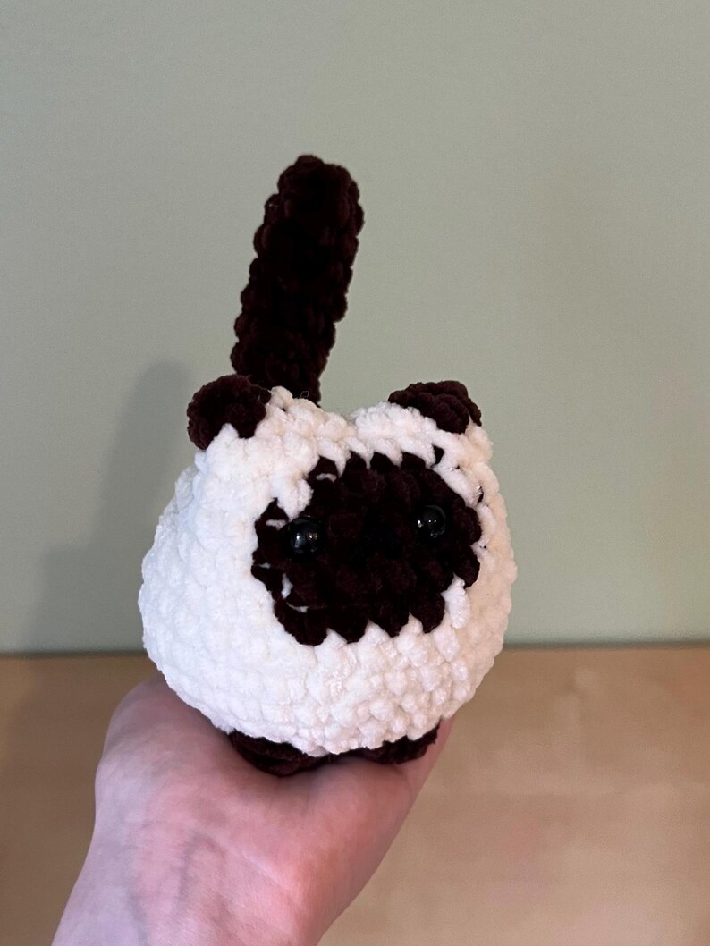 May include: A handmade crochet cat toy in white and brown. The cat has dark brown markings around the eyes, ears, and paws, with a matching tail. The toy is round and fluffy, with black safety eyes.
