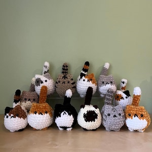 May include: A collection of handmade crochet cat butt ornaments in various colors and patterns. Each cat butt features a fluffy tail and a round body. The color palette includes white, brown, black, gray, and orange.