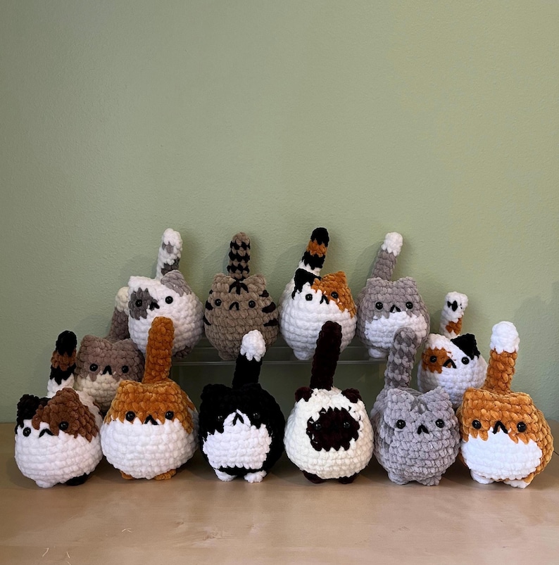 May include: A collection of handmade crochet cat butt ornaments in various colors and patterns. Each cat butt features a fluffy tail and a round body. The color palette includes white, brown, black, gray, and orange.