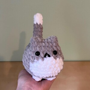 May include: A handmade, gray and white crochet cat plushie with black button eyes and a small, upturned nose. The cat has a fluffy, rounded body and a tail that is raised. The plushie is a cute, whimsical design.