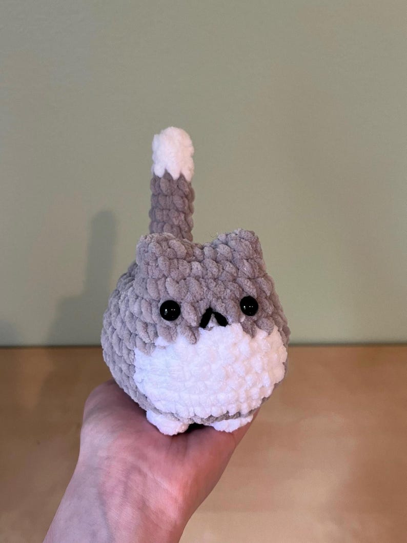 May include: A handmade, gray and white crochet cat plushie with black button eyes and a small, upturned nose. The cat has a fluffy, rounded body and a tail that is raised. The plushie is a cute, whimsical design.