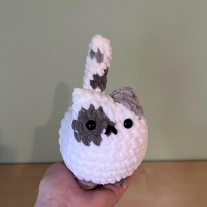 May include: A handmade, white crocheted cat with gray spots and a tail. The cat has black button eyes and a small, gray ear. The plush toy is round and soft, perfect for a child's room or as a decorative item.