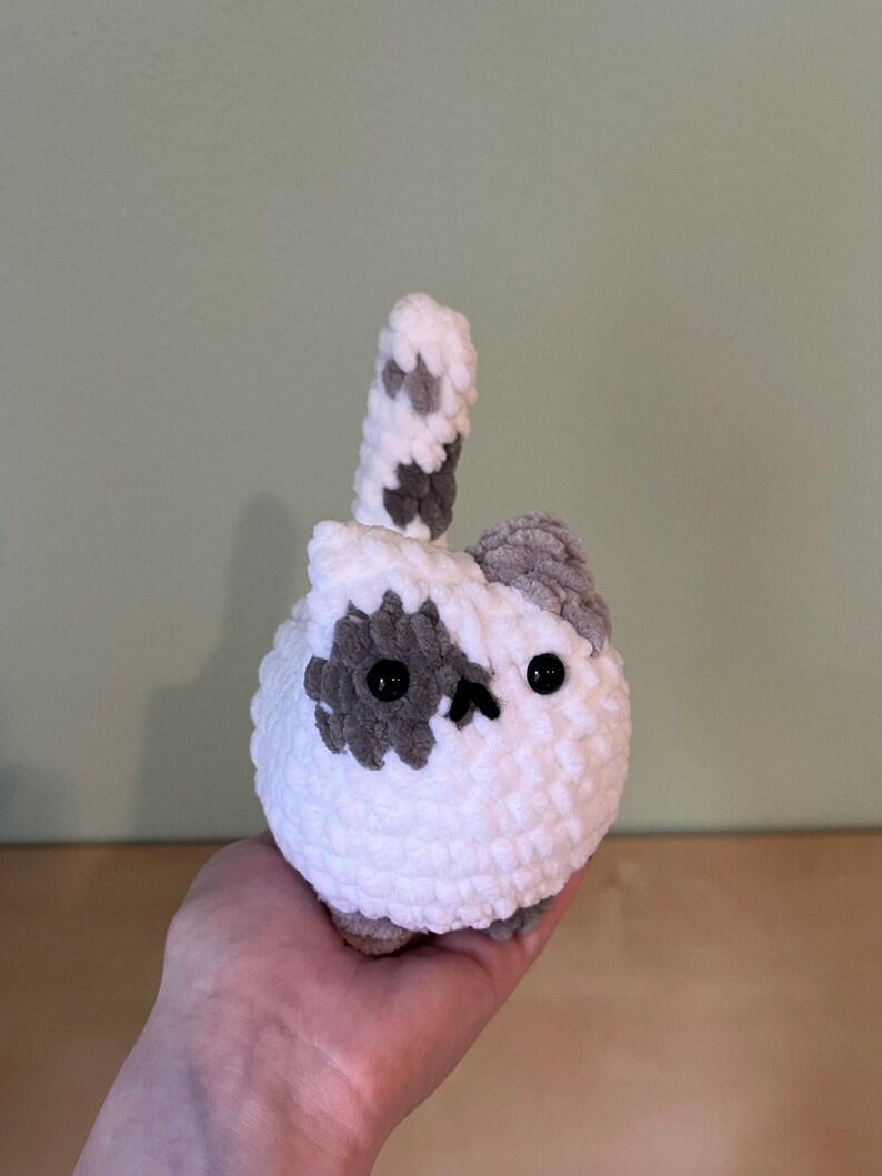 May include: A handmade, white crocheted cat with gray spots and a tail. The cat has black button eyes and a small, gray ear. The plush toy is round and soft, perfect for a child's room or as a decorative item.