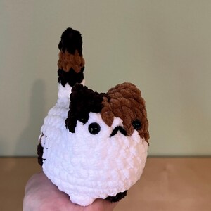 May include: A handmade, plush cat toy with a white body, and patches of brown and black. The cat has small black eyes and a tail with brown, black, and white stripes. The toy is held in a person's hand.