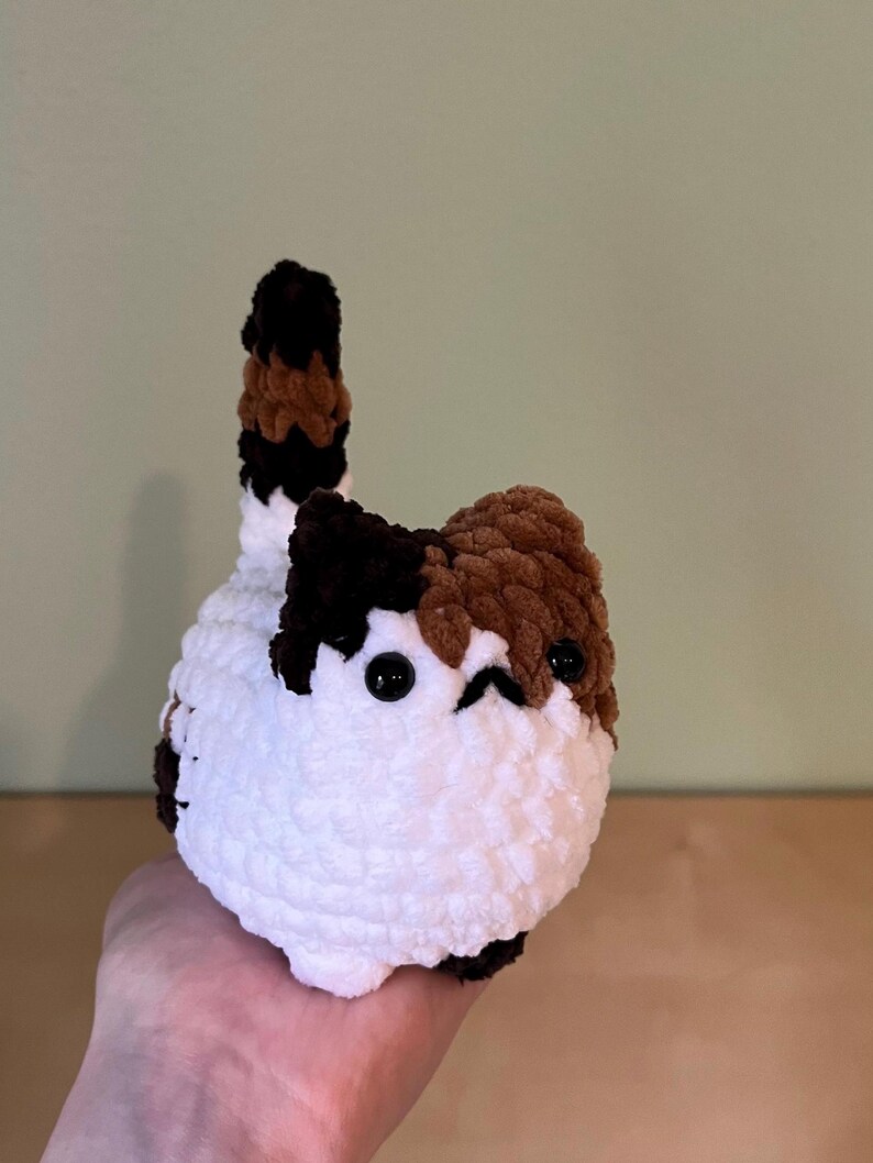 May include: A handmade, plush cat toy with a white body, and patches of brown and black. The cat has small black eyes and a tail with brown, black, and white stripes. The toy is held in a person's hand.