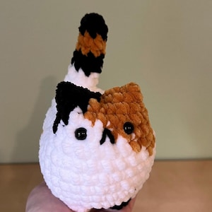 May include: A handmade crochet cat with a white body, and patches of brown and black. The cat has black eyes and a tail with black, brown, and white stripes. The cat is held in a person's hand.