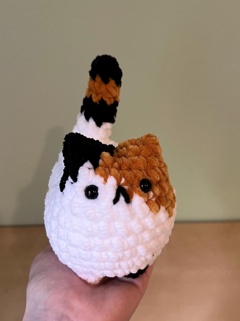 May include: A handmade crochet cat with a white body, and patches of brown and black. The cat has black eyes and a tail with black, brown, and white stripes. The cat is held in a person's hand.