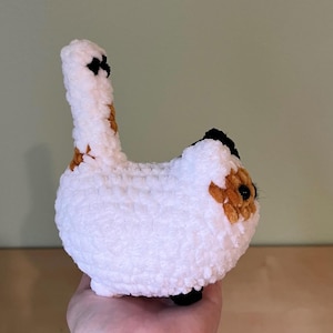May include: A handmade, white crochet cat with black and brown markings. The cat has a long tail and is held in a person's hand. The cat's body is round and fluffy, with small black feet.