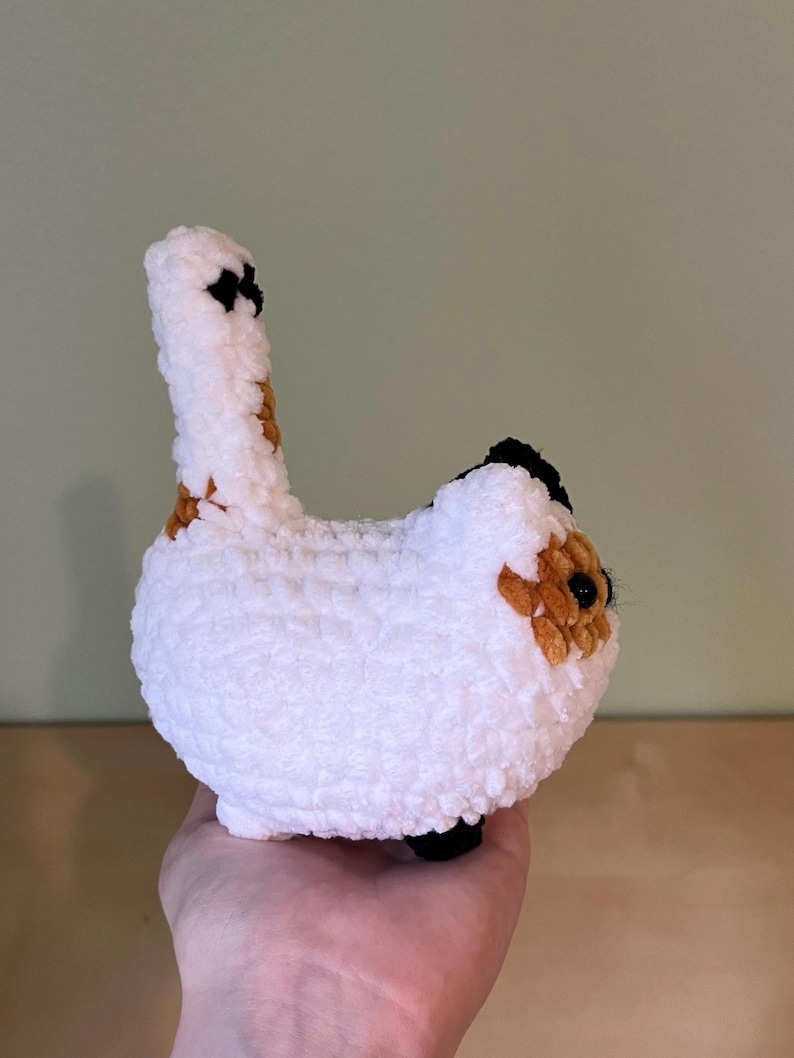 May include: A handmade, white crochet cat with black and brown markings. The cat has a long tail and is held in a person's hand. The cat's body is round and fluffy, with small black feet.