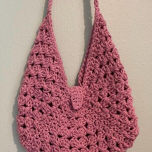 Puede incluir: Bolso de ganchillo rosa hecho a mano con correa para el hombro. El bolso presenta un patr&oacute;n de tejido abierto y texturizado y un peque&ntilde;o cierre de solapa. Cuelga contra una pared azul claro.